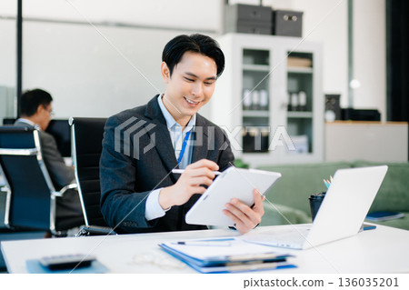 Confident Asian businessman using tablet and laptop in bright modern office, representing digital work, innovation Confident Asian businessman using tablet and laptop in bright modern office, representing digital work, innovation 136035201