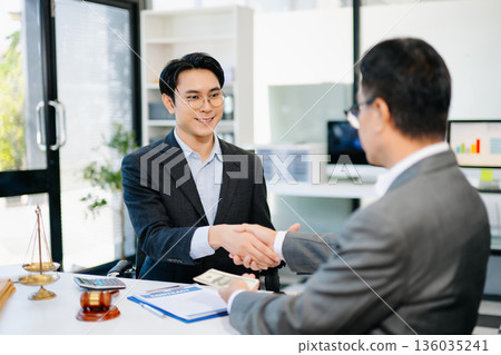 Lawyer and client shaking hands after contract signing with money and gavel on desk. Ideal for business, finance, partnership, or legal themes. Lawyer and client shaking hands after contract signing with money and gavel on desk. Ideal for business, finance, partnership, or legal themes. 136035241