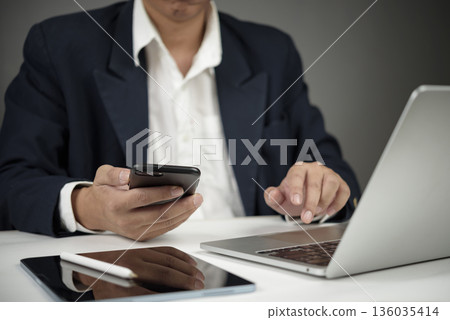 Businessman in suit working with smartphone and laptop at desk, modern office concept with mobile technology and digital communication tools. Businessman in suit working with smartphone and laptop at desk, modern office concept with mobile technology and digital communication tools. 136035414