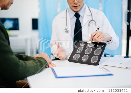 Doctor showing brain MRI results to patient during consultation in modern clinic. Perfect for healthcare, diagnosis, neurology, or medical treatment themes. Doctor showing brain MRI results to patient during consultation in modern clinic. Perfect for healthcare, diagnosis, neurology, or medical treatment themes. 136035421