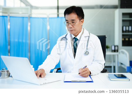 Asian doctor working on laptop and writing notes in modern clinic. Ideal for healthcare, diagnosis, medical research, and professional consultation themes. Asian doctor working on laptop and writing notes in modern clinic. Ideal for healthcare, diagnosis, medical research, and professional consultation themes. 136035513