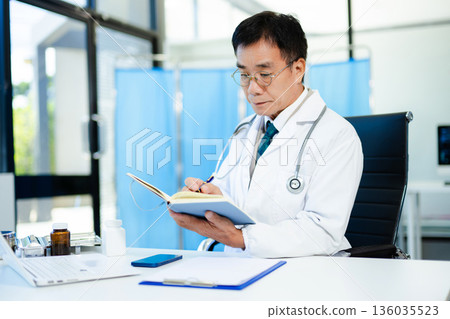 Asian doctor working on laptop and writing notes in modern clinic. Ideal for healthcare, diagnosis, medical research, and professional consultation themes. Asian doctor working on laptop and writing notes in modern clinic. Ideal for healthcare, diagnosis, medical research, and professional consultation themes. 136035523