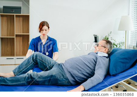 A nurse in blue scrubs guides a senior patient through leg mobility exercises on a therapy bed, showcasing real people A nurse in blue scrubs guides a senior patient through leg mobility exercises on a therapy bed, showcasing real people 136035868