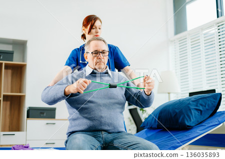 Home physiotherapy: Nurse in blue scrubs guides an elderly patient through resistance band exercises, showcasing real people, mobility Home physiotherapy: Nurse in blue scrubs guides an elderly patient through resistance band exercises, showcasing real people, mobility 136035893