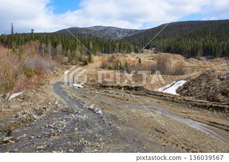 Muddy dirt road with a stream flowing through it, flanked by hills with logging clearcuts and a green coniferous forest Muddy dirt road with a stream flowing through it, flanked by hills with logging clearcuts and a green coniferous forest 136039567
