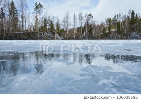 Melting ice on a lake surface with reflections of bare trees and evergreen forest in Ruskeala Mountain Park. Spring nature thaw concept. 136039580