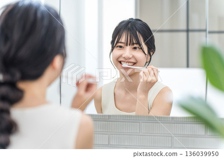 一位年輕的亞洲女性在浴室裡對著鏡子刷牙。 136039950