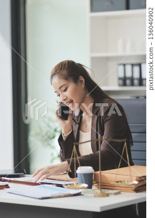 Confident Lawyer Working at Desk: A young Asian female lawyer confidently works on her laptop, surrounded by legal symbols. She exudes professionalism and competence in her modern office setting. Confident Lawyer Working at Desk: A young Asian female lawyer confidently works on her laptop, surrounded by legal symbols. She exudes professionalism and competence in her modern office setting. 136040902