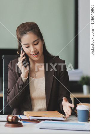 Confident Lawyer Working at Desk: A young Asian female lawyer confidently works on her laptop, surrounded by legal symbols. She exudes professionalism and competence in her modern office setting. 136040903
