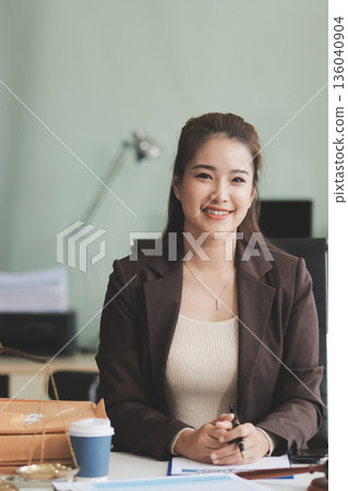 Confident Lawyer Working at Desk: A young Asian female lawyer confidently works on her laptop, surrounded by legal symbols. She exudes professionalism and competence in her modern office setting. Confident Lawyer Working at Desk: A young Asian female lawyer confidently works on her laptop, surrounded by legal symbols. She exudes professionalism and competence in her modern office setting. 136040904