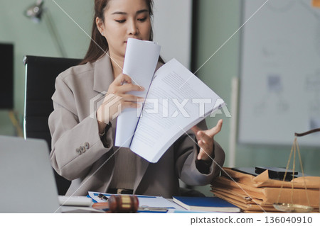 Confident Lawyer Working at Desk: A young Asian female lawyer confidently works on her laptop, surrounded by legal symbols. She exudes professionalism and competence in her modern office setting. Confident Lawyer Working at Desk: A young Asian female lawyer confidently works on her laptop, surrounded by legal symbols. She exudes professionalism and competence in her modern office setting. 136040910