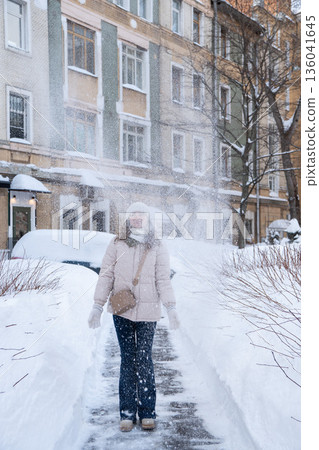 Young woman walking along a snowy residential sidewalk while snow falls around her, wearing light winter jacket, knit hat and crossbody bag Young woman walking along a snowy residential sidewalk while snow falls around her, wearing light winter jacket, knit hat and crossbody bag 136041645