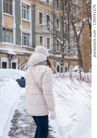 Woman in a light winter jacket walks along a snowy residential sidewalk, urban winter lifestyle with calm mood and neutral tones Woman in a light winter jacket walks along a snowy residential sidewalk, urban winter lifestyle with calm mood and neutral tones 136041876