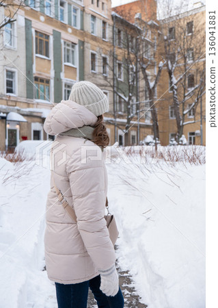 Woman in a light winter jacket walks along a snowy residential sidewalk, urban winter lifestyle with calm mood and neutral tones Woman in a light winter jacket walks along a snowy residential sidewalk, urban winter lifestyle with calm mood and neutral tones 136041881
