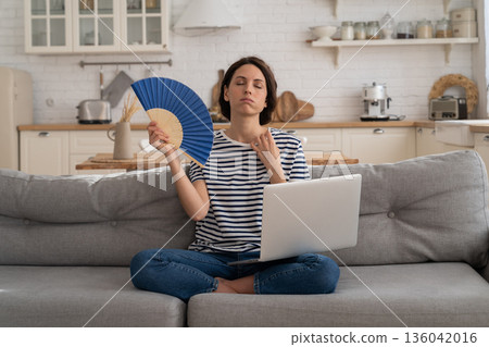 Young woman suffers from heatstroke flat without air-conditioner waving fan sitting on couch at home 136042016