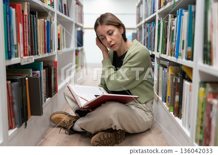 Pretty concentrated adult female sit on floor between bookshelves, reading interesting book on knees 136042033