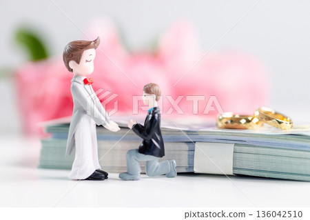 Two groom figurines, one kneeling to propose, stand beside stack of banknotes with gold wedding rings and soft pink flowers in background, symbolizing inclusive love and equality. inclusive love Two groom figurines, one kneeling to propose, stand beside stack of banknotes with gold wedding rings and soft pink flowers in background, symbolizing inclusive love and equality. inclusive love 136042510