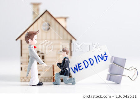 Two groom figurines, one kneeling to propose, stand before miniature house with a WEDDING card and binder clip on clean white background with copy space, expressing inclusive commitment and equality. Two groom figurines, one kneeling to propose, stand before miniature house with a WEDDING card and binder clip on clean white background with copy space, expressing inclusive commitment and equality. 136042511