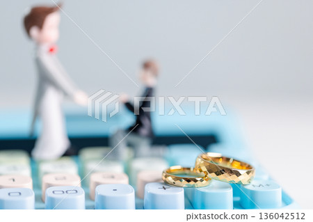 Two gold wedding rings on blue calculator keys with blurred groom figurines in background on white copy space, highlighting budgeting, savings and inclusive wedding planning. budgeting, savings Two gold wedding rings on blue calculator keys with blurred groom figurines in background on white copy space, highlighting budgeting, savings and inclusive wedding planning. budgeting, savings 136042512