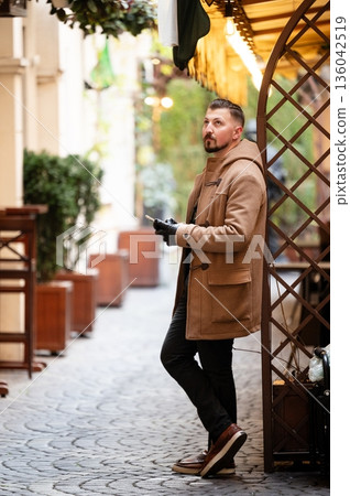 Stylish man stands thoughtfully in a quaint alleyway cafe as autumn leaves create a warm backdrop in a charming urban setting Stylish man stands thoughtfully in a quaint alleyway cafe as autumn leaves create a warm backdrop in a charming urban setting 136042519