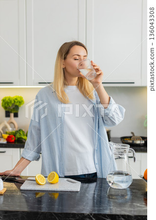 Young woman squeezing lemon into water tumbler in kitchen for fresh vitamin hydration morning habit 136043308