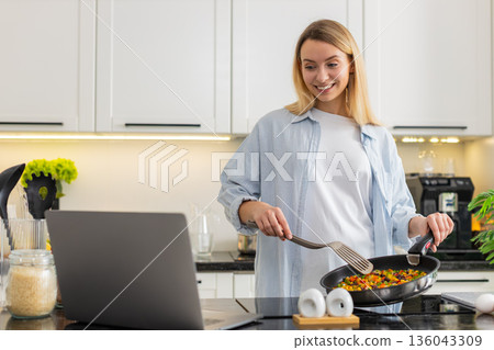 Young woman cooking vegetables on electric stove while watching keto recipe on laptop in own kitchen Young woman cooking vegetables on electric stove while watching keto recipe on laptop in own kitchen 136043309