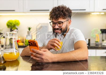 Arabian young adult guy drinking fresh orange juice in kitchen at home reading news on smartphone Arabian young adult guy drinking fresh orange juice in kitchen at home reading news on smartphone 136043328