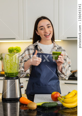 Young woman pours green smoothie into glass after blending fruits, spinach, healthy breakfast drink 136043341