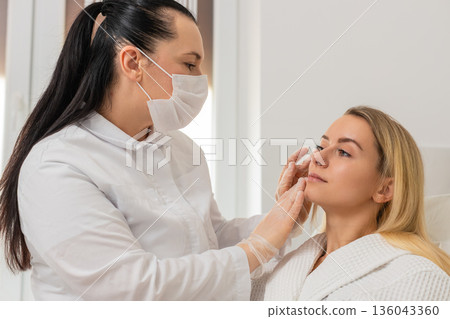 Cosmetologist in mask checking facial skin of young woman client before cosmetic treatment in clinic 136043360