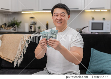 Asian man counts cash dollars smiles makes money sign and plans invest purchase bonus salary savings Asian man counts cash dollars smiles makes money sign and plans invest purchase bonus salary savings 136043466