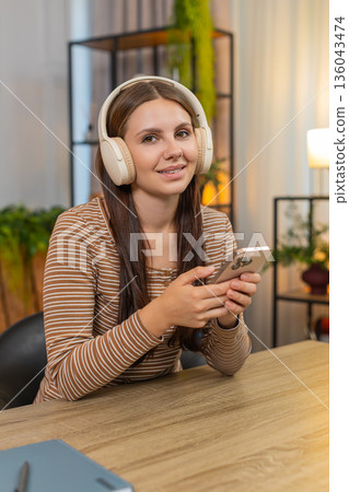 Young woman in headphones listens to work podcast saves ideas to notes on smartphone while focused Young woman in headphones listens to work podcast saves ideas to notes on smartphone while focused 136043474