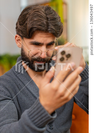 Indian man with eyeglasses squints at smartphone screen tries to read small text poor vision focus 136043497