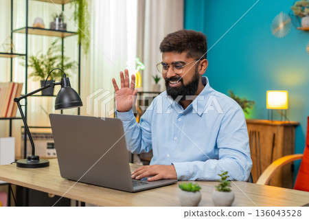 Indian man waves greeting speaks via laptop webcam in video call smiles during online conference web 136043528