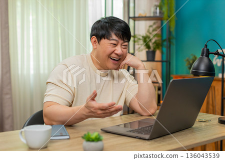 Asian man smiling watching nostalgic video on laptop, resting head on hand, enjoying warm memories 136043539