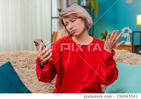 Young woman receiving bad news on smartphone screen, looking upset, disappointed shaking head sadly Young woman receiving bad news on smartphone screen, looking upset, disappointed shaking head sadly 136043541