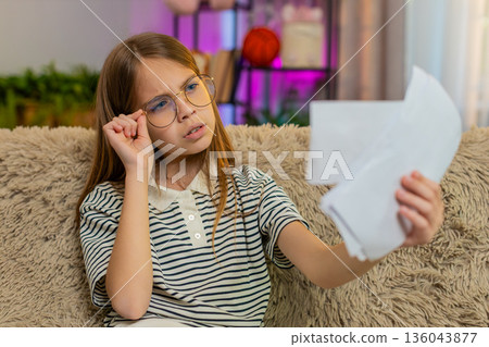 Child girl kid pretending to read documents in adult glasses sitting on home sofa at living room 136043877