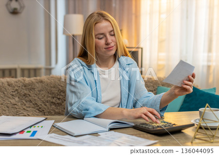 Young woman writing budget at home, table workspace, documents, calculator focused plan calm routine Young woman writing budget at home, table workspace, documents, calculator focused plan calm routine 136044095