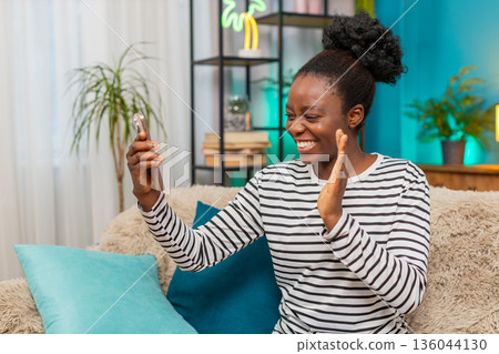 African American woman smiling waving smartphone hand gesture greeting happy indoors sofa African American woman smiling waving smartphone hand gesture greeting happy indoors sofa 136044130