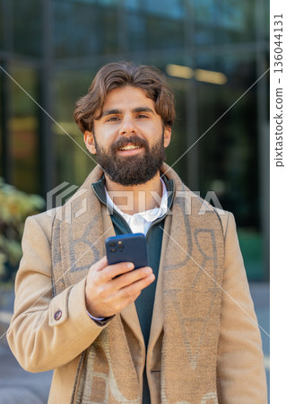 Happy young business man using smartphone and looking at camera while standing in downtown street Happy young business man using smartphone and looking at camera while standing in downtown street 136044131