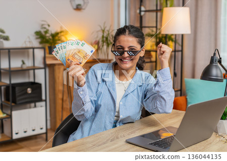 Young woman freelancer putting on party glasses holding fan of cash sitting at home office table Young woman freelancer putting on party glasses holding fan of cash sitting at home office table 136044135