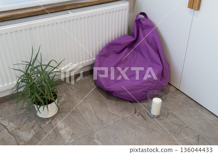 A purple bean bag is placed in the corner of a well-lit room. A small plant is in a pot nearby along with a candle on the floor A purple bean bag is placed in the corner of a well-lit room. A small plant is in a pot nearby along with a candle on the floor 136044433