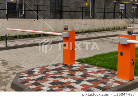 Two vibrant orange barriers stand guard at a contemporary parking lot entrance, surrounded by neatly arranged bricks and green grass 136044453