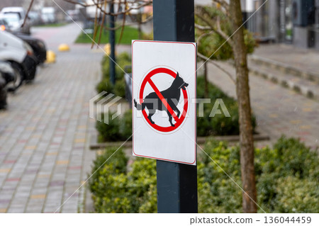 A clear no dog walking sign stands tall on a post in a city area, surrounded by neatly paved roads and vibrant greenery A clear no dog walking sign stands tall on a post in a city area, surrounded by neatly paved roads and vibrant greenery 136044459