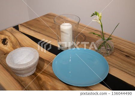 A beautiful display of a blue plate, a glass candle holder with a white candle, and a delicate flower in a vase, all on a wooden table 136044490
