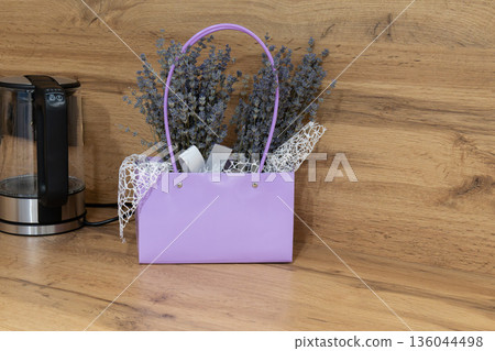 A charming lavender bag holds fragrant lavender flowers, adding a pop of color to the warm wooden counter beside a sleek kettle A charming lavender bag holds fragrant lavender flowers, adding a pop of color to the warm wooden counter beside a sleek kettle 136044498