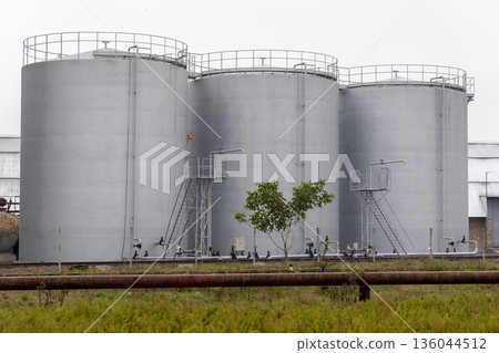 Silver storage tanks tower against a gray sky, surrounded by green grass and industry, showcasing human innovation and nature Silver storage tanks tower against a gray sky, surrounded by green grass and industry, showcasing human innovation and nature 136044512