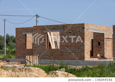 Bright sunlight highlights the vibrant bricks of a nearly finished home, surrounded by lush greenery and soft soil Bright sunlight highlights the vibrant bricks of a nearly finished home, surrounded by lush greenery and soft soil 136044548
