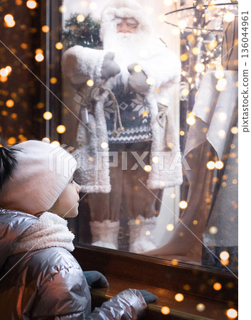 Child gazes at Santa in a window display filled with warm lights and festive wonder 136044961