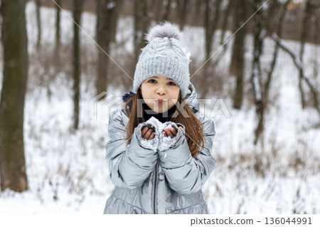 Girl in a silver puffer jacket blowing snow in a snowy forest during winter 136044991