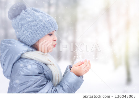 Young child in a blue winter coat blows snow from gloved hands outdoors 136045003